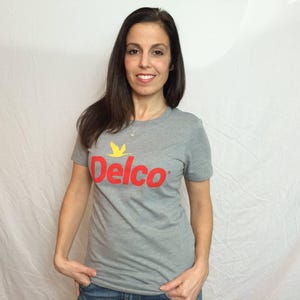 Peut inclure: Un t-shirt gris avec un logo rouge et jaune qui dit "Delco".