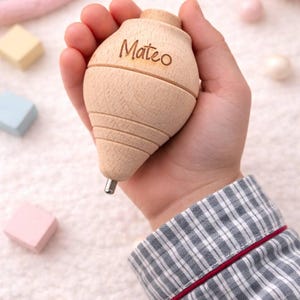 Può includere: Una trottola di legno chiaro, incisa con il nome "Mateo". La trottola presenta un design scanalato e una punta metallica. L'immagine mostra un primo piano della trottola tenuta in mano.