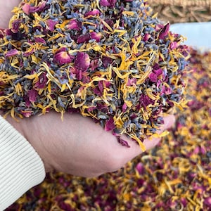 May include: A handful of dried flower petals in shades of purple, yellow, and pink. The mix includes rose petals, lavender buds, and marigold petals. The petals are held in a cupped hand, with more petals scattered below.