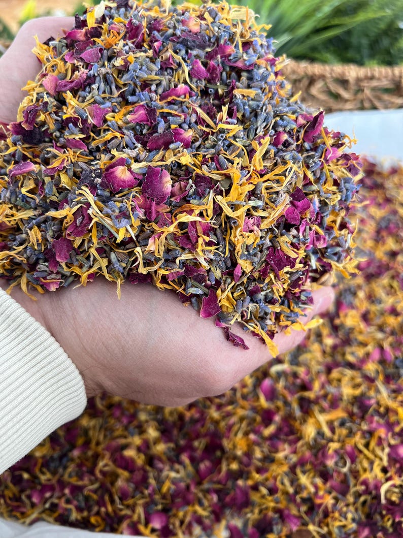 May include: A handful of dried flower petals in shades of purple, yellow, and pink. The mix includes rose petals, lavender buds, and marigold petals. The petals are held in a cupped hand, with more petals scattered below.