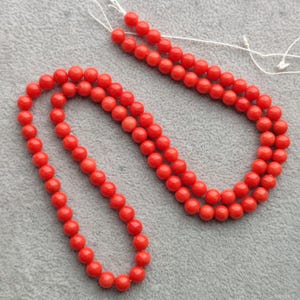 Puede incluir: Un collar de cuentas rojas vibrantes, ensartadas en hilo blanco, descansa sobre una superficie gris. Las cuentas son redondas y de tamaño uniforme, creando una línea continua. Probablemente un accesorio de moda.