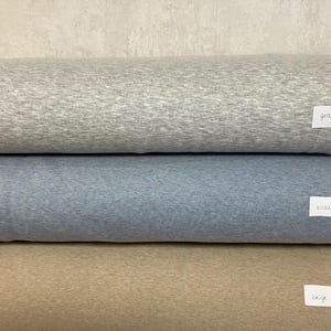 Könnte beinhalten: Drei Stoffrollen in Grau-, Blau- und Beigetönen. Jede Rolle hat ein kleines weißes Etikett mit einem Farbnamen. Der Stoff scheint ein weiches, strukturiertes Material zu sein.