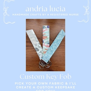Puede incluir: Tres llaveros de tela con herrajes metálicos. Uno presenta un patrón floral gris, otro un estampado acuático colorido con peces y el tercero un patrón de espirales azul claro. El texto dice andria lucia HANDMADE CRAFTS BY A REGISTERED NURSE Custom Key Fob PICK YOUR OWN FABRIC & I’LL CREATE A CUSTOM KEEPSAKE FOR YOU!