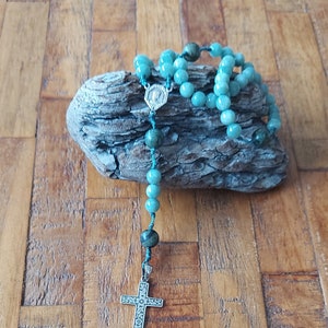 Peut inclure: Un chapelet en pierres précieuses bleu turquoise et vert avec un pendentif en croix argentée. Le chapelet repose sur un morceau de bois flotté.