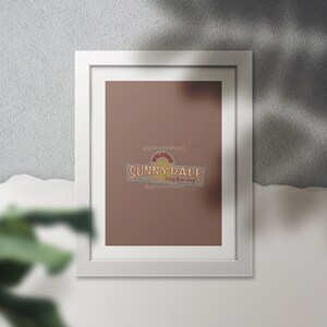 Op de afbeelding: Een witte ingelijste print met een bruine achtergrond en een retro-logo dat "Sunnydale" zegt met de tekst "Welcome" en "Enjoy Your Stay" in een cirkel boven en onder het woord "Sunnydale". Het logo is in geel, rood en oranje.