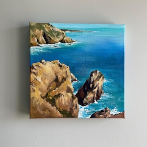 Könnte beinhalten: Quadratische Leinwandmalerei einer Küstenlandschaft. Das Kunstwerk zeigt einen lebendigen blauen Ozean, der auf zerklüftete, braune Klippen trifft. Der Himmel ist hellblau und das Gemälde fängt die natürliche Schönheit der Küste ein.