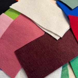 May include: A collection of colorful felt rectangles in various shades. The assortment includes white, pink, burgundy, green, and blue pieces. The felt appears to be soft and evenly textured, suitable for crafts and DIY projects.
