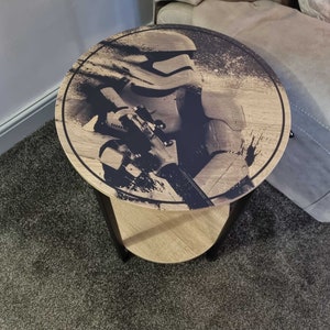 May include: A round wooden side table with a black metal frame. The table top features a black and white graphic of a Star Wars Stormtrooper holding a blaster rifle.