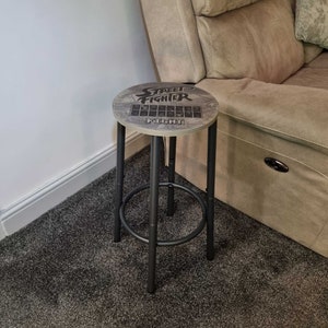 May include: A black metal stool with a round wooden top. The top has a wood grain pattern and the words "Street Fighter" printed on it. The stool is sitting on a gray carpet.
