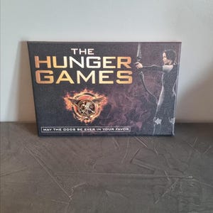 以下が含まれることがあります： 「THE HUNGER GAMES」の文字が金色で書かれた長方形のキャンバスプリント。炎のエンブレムの上にあります。右側には弓矢を持った人物がいます。「MAY THE ODDS BE EVER IN YOUR FAVOR」というフレーズが一番下にあります。