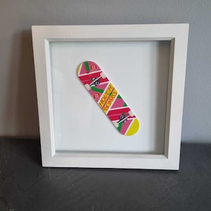 Puede incluir: Una imagen enmarcada en blanco con una patineta rosa, verde y amarilla en su interior. La patineta tiene las palabras "Hover Board" impresas en ella.