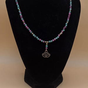 Puede incluir: Un collar de cuentas con pequeñas perlas facetadas que alternan entre turquesa y magenta, con espaciadores dorados. Una sección central desciende hacia un pequeño colgante de metal color bronce con forma de flor de loto.