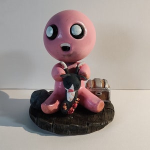 May include: A pink figurine of a character with large eyes and a wide mouth, holding a black and white cat figurine. The character is sitting on a wooden base with a small treasure chest.