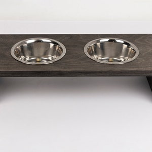May include: A raised pet feeder with two stainless steel bowls set into a dark gray wooden frame. The frame has a rectangular shape with a minimalist design, providing an elevated feeding solution for pets.