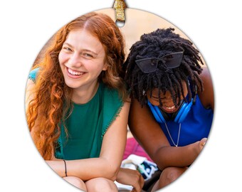 May include: Two friends, one with red hair and wearing a green shirt, and the other with dark hair and wearing a blue shirt, are laughing together. They are sitting on a bench outdoors.
