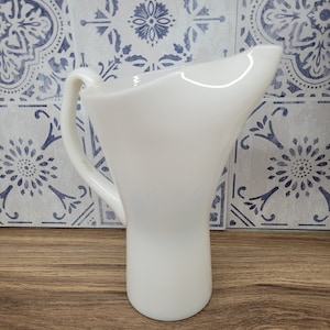 May include: A white pitcher with a curved handle and a flared top. The jug is made of a smooth, opaque material and sits on a wooden surface. The background features a blue and white patterned tile.