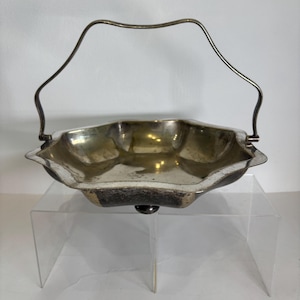 May include: A silver-toned metal basket with a hexagonal, scalloped rim and a single arched handle. The interior surface features a reflective, tarnished finish, while the exterior base is supported by small, dark, rounded feet.