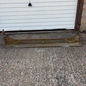 May include: A long, rectangular brass fireplace fender featuring ornate decorative detailing, including floral swags, vertical slats, and finials at each end. The metal surface displays a weathered patina with visible signs of age and oxidation.