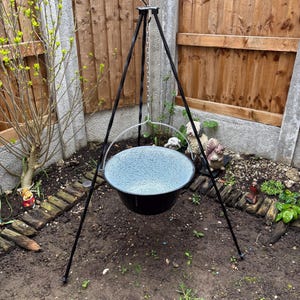 May include: A black metal tripod stand with a light-coloured wooden handle at the top, supporting a round, deep cooking pot. The pot features a black exterior and a speckled, light blue and white interior, suspended by a metal chain and handle.