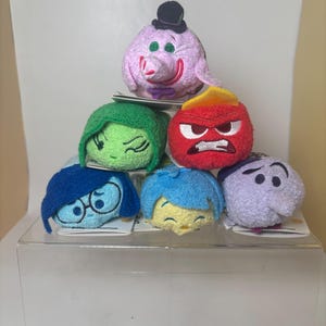 May include: A pyramid of plush toys featuring characters from the movie Inside Out. The toys include characters like Anger, Sadness, Joy, and Fear. Each plush has a unique design and color scheme, stacked on a clear display case.