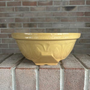 May include: A mustard yellow ceramic mixing bowl with a wide, flat rim and a decorative embossed pattern around the exterior. The bowl features a sturdy base and a smooth, glossy finish, showcasing a classic design with a wide opening.