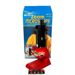 Puede incluir: Una caja azul con las palabras "Zoom Microscope" y "Age from 5" impresas. Un microscopio rojo con un ocular negro está frente a la caja. Una diapositiva con muestras de insectos está en el microscopio.