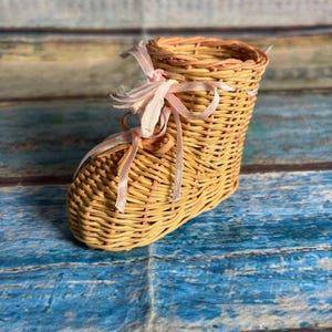 May include: A small, boot-shaped container crafted from woven light brown wicker. The boot features a decorative, pale pink ribbon tied in a bow at the front, serving as laces. The structure is hollow, with a circular opening at the top.