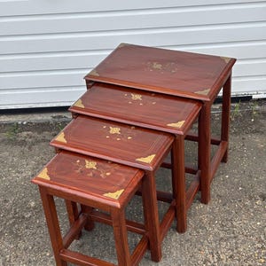 May include: A set of three nesting tables in a deep reddish-brown colour. Each table has a square top with gold-coloured corner detailing and floral inlay. The tables are designed to fit inside each other, with varying sizes.