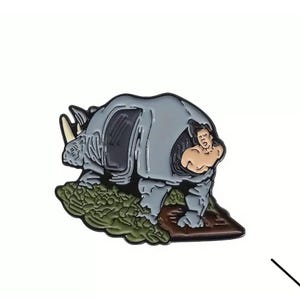 May include: A grey rhinoceros pin with a man riding on its back. The rhinoceros is standing on a patch of green foliage. The man is wearing a white shirt and has brown hair.