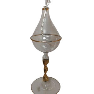 May include: A clear glass oil lamp with a gold-coloured rim and a twisted gold-coloured stem. The lamp has a teardrop-shaped reservoir and a round base. A wick extends from the top.