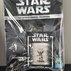 Op de afbeelding: Een verpakte Star Wars-beeldjesserie, met een Watto-figuur. De verpakking bevat het Star Wars-logo, het nummer 50 en de tekst "The Official Figurine Collection". Het beeldje zit in een doorzichtige plastic hoes.