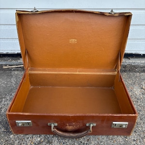 May include: An open brown leather suitcase with a tan interior lining. The suitcase features a central handle, two silver-toned metal latches on the front, and a small gold-coloured logo printed on the inside of the lid.
