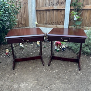 May include: Two matching side tables with a dark cherry wood finish. Each table features a rectangular top, a single drawer with a curved brass-toned metal handle, and a sturdy base with two vertical supports connected by a horizontal stretcher.