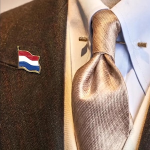 Op de afbeelding: Een goudkleurige speld met de Nederlandse vlag, met rode, witte en blauwe strepen. De speld is bevestigd aan een bruine tweed blazer, gecombineerd met een lichtblauw overhemd en een beige stropdas.