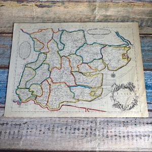 May include: An antique-style paper map of Essex, England, featuring hand-colored borders in red, blue, and yellow. The map includes detailed cartographic markings, a decorative title cartouche in the bottom right corner, and a compass rose.