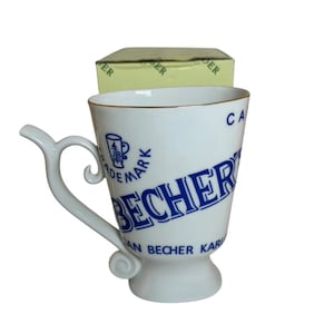 以下が含まれることがあります： 青いロゴと文字が入った白いセラミックマグカップ。「BECHER」は様式化されたフォントです。マグカップはユニークなハンドルデザインと金の縁取りが特徴です。マグカップの後ろに小さな箱が見えます。