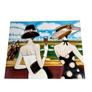 May include: A ceramic tile depicting two women at a racecourse. One is in a black hat and white dress, holding a glass of red wine. The other wears a beige hat and a black polka dot dress. The background shows a race track with horses and jockeys.