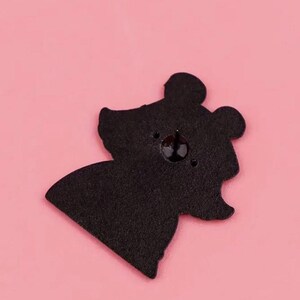 May include: Black enamel pin featuring a cartoon bear with a simple design. The bear has a round head with two small ears and a small nose. The pin has a silver metal backing.