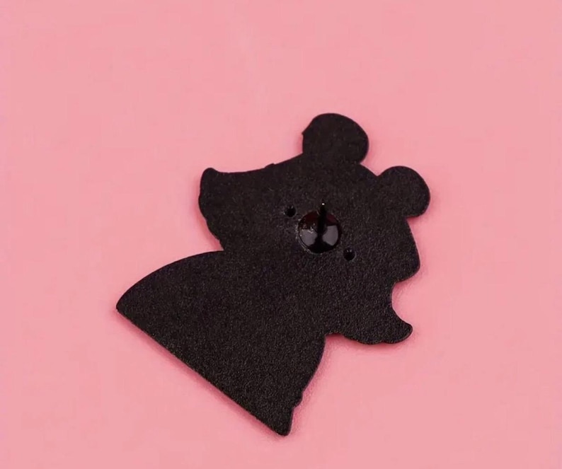May include: Black enamel pin featuring a cartoon bear with a simple design. The bear has a round head with two small ears and a small nose. The pin has a silver metal backing.