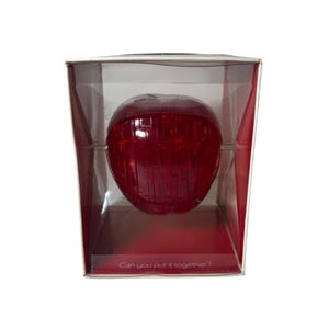 May include: A red apple-shaped puzzle inside a clear plastic box. The box has a red base and the text "Can you put it together?" is printed on the base.