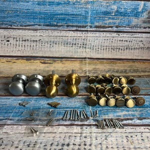 May include: A selection of door knobs and associated hardware in silver, brass, and bronze finishes. The knobs are round, with some featuring decorative elements. Screws and other fixings are also present.