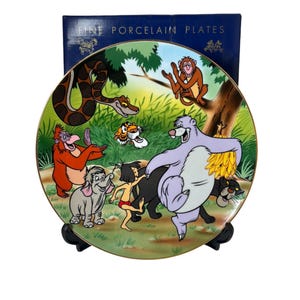 Può includere: Un piatto decorativo in porcellana con personaggi de Il libro della giungla. Il piatto ha un bordo dorato e un'illustrazione colorata di Baloo, Bagheera, Mowgli e altri personaggi in un ambiente della giungla. Il piatto è esposto con una scatola blu scuro che recita "FINE PORCELAIN PLATES."