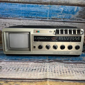 May include: A silver Lloytron portable colour TV, radio, and cassette tape recorder featuring a small screen on the left, a radio frequency display with multiple bands, six black control knobs, and a row of black push-button switches on the top right.