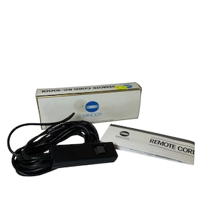 May include: A black Minolta remote cord with a black control box and a white box with the text "REMOTE CORD RC-1000L". A small white booklet with the text "REMOTE CORD" is also included.