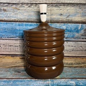 May include: A glossy brown ceramic table lamp base with a cylindrical, ribbed, stacked-ring design. The lamp features a white plastic light bulb socket extending from the top centre and a thin white electrical cord extending from the base.