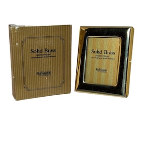 May include: A solid brass photo frame with a gold finish, next to its original cardboard box. The frame has a rectangular shape with rounded corners and the text "Solid Brass Photo Frame" printed on the front. The box also has the same text.
