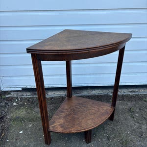 May include: A dark brown wooden corner table with a triangular top and a lower shelf. The table has three legs and a simple design. The top and shelf are semi-circular. The table is made of wood.