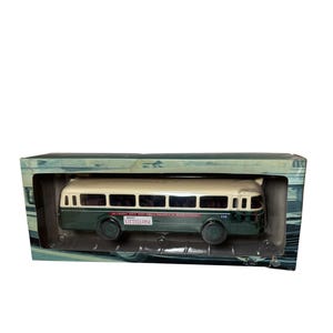 May include: A vintage toy bus model in a display box. The bus is two-toned, with a cream-coloured roof and a dark green body. The bus has black wheels and several windows. The box has a green and blue design.