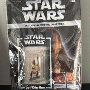 Puede incluir: Colección de figuras de Star Wars empaquetada con Qui-Gon Jinn. El paquete incluye una pequeña figura del personaje sosteniendo un sable de luz, con el logo de Star Wars y el texto "The Official Figurine Collection". El número 42 es visible.