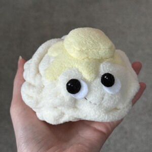 Puede incluir: Un peluche con forma de coliflor, con una cara sonriente y grandes ojos negros. El juguete es principalmente blanquecino con una parte superior de color amarillo claro, sostenido en una mano.